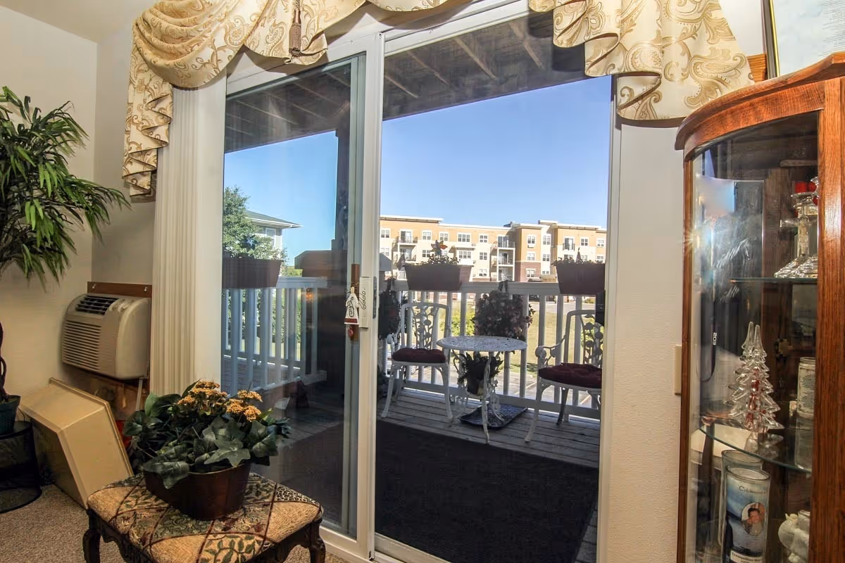 View of a small balcony with a white metal table and two chairs with red cushions, seen through a glass sliding door from inside a room. The room has a potted plant on a small table, an air conditioning unit on the wall, and a wooden display cabinet with glass shelves containing decorative items. Outside, there are flower pots on the balcony railing and a multi-story apartment building in the background under a clear blue sky.