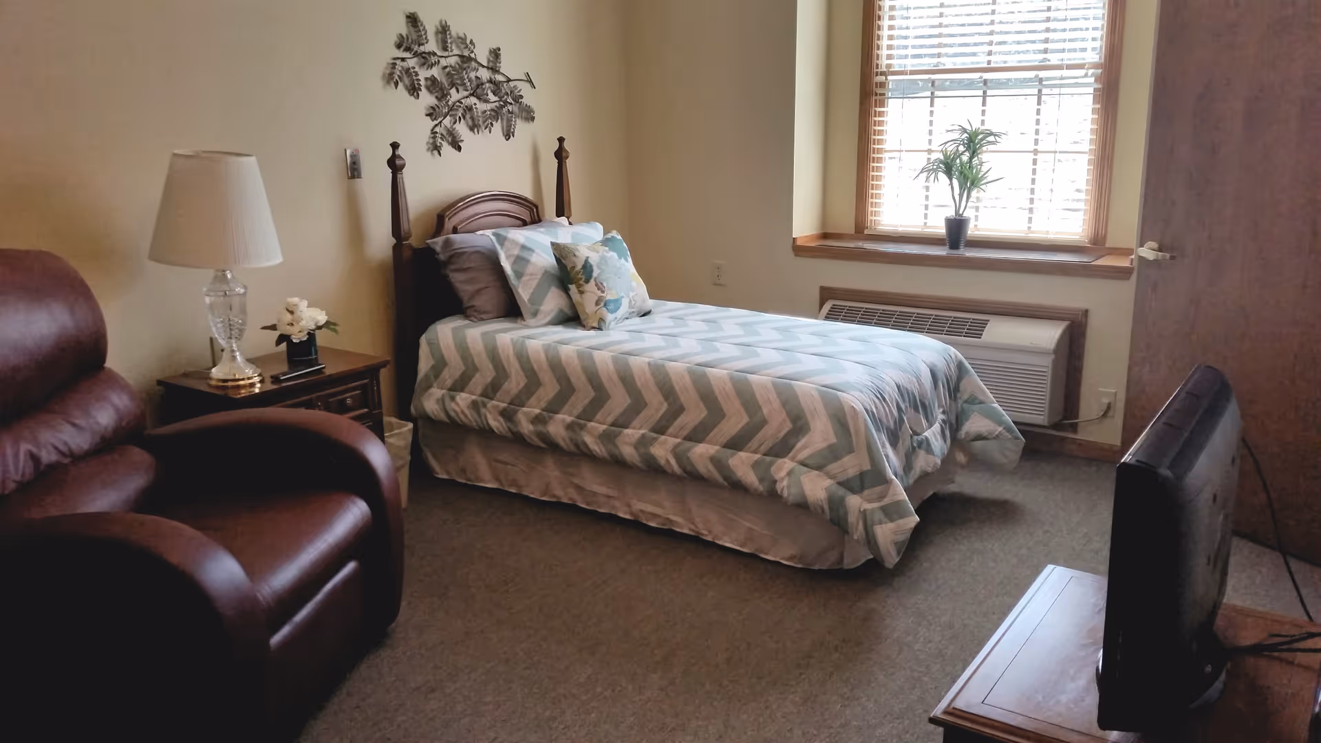A cozy bedroom with a single bed covered in a chevron-patterned comforter and multiple pillows. Next to the bed is a wooden nightstand with a lamp and a small flower arrangement. A brown leather recliner chair is positioned near the nightstand. A window with blinds and a small potted plant on the windowsill lets in natural light. A television is placed on a wooden stand opposite the bed.