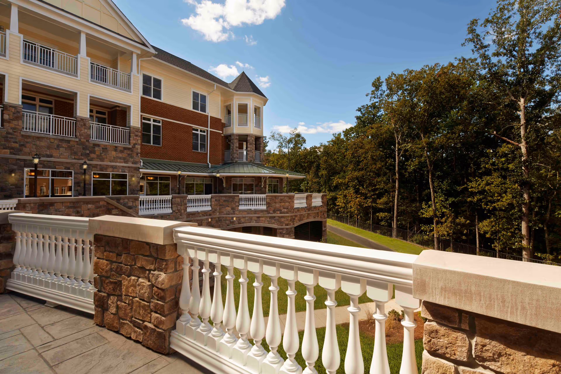 View of a multi-story senior living facility building with stone and brick exterior, white railings on balconies and terraces, overlooking a green lawn and trees under a partly cloudy sky.