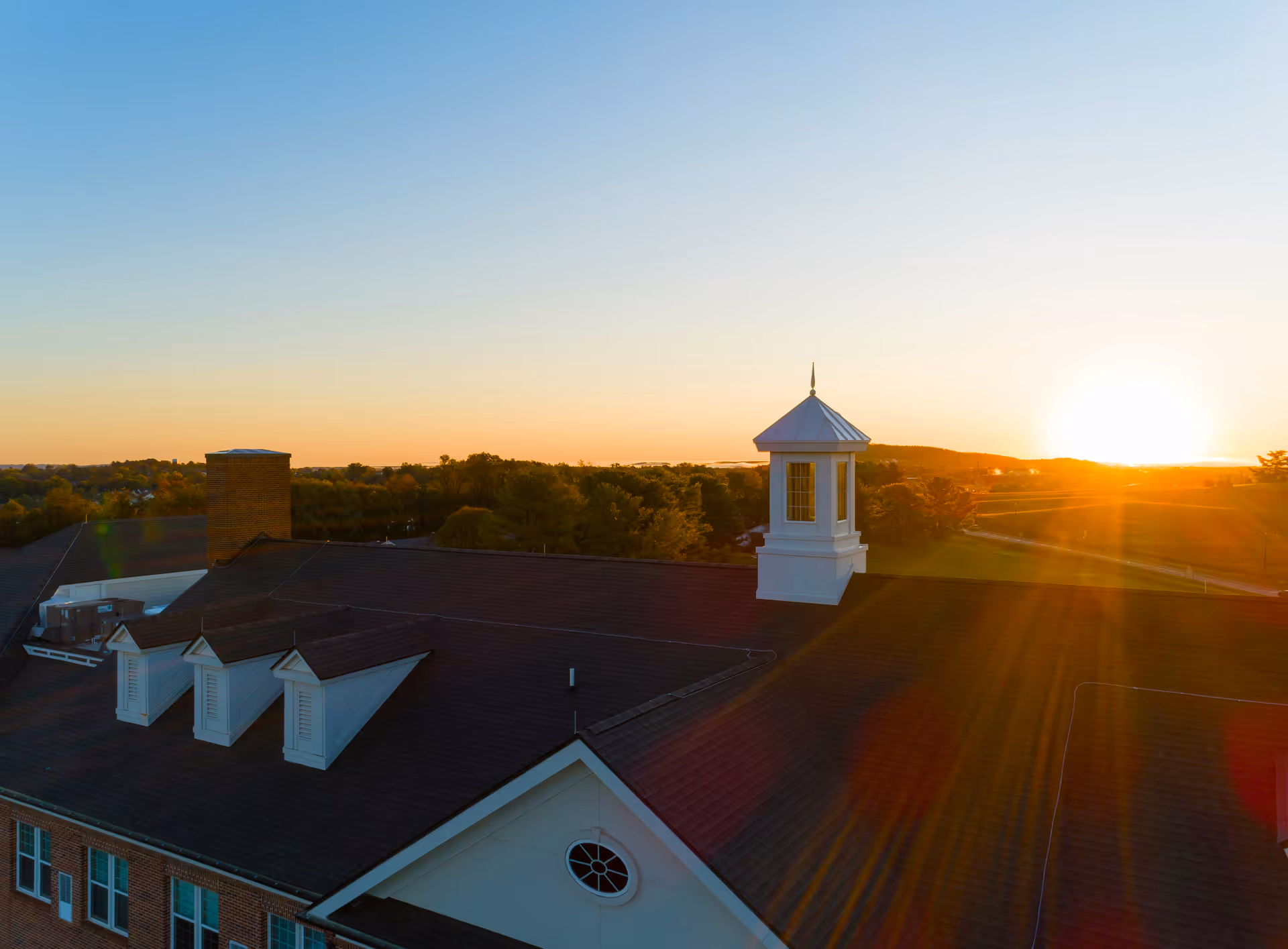 Aerial view of The Culpeper building roof at sunset with a white cupola and chimney, surrounded by trees and open landscape under a clear sky.