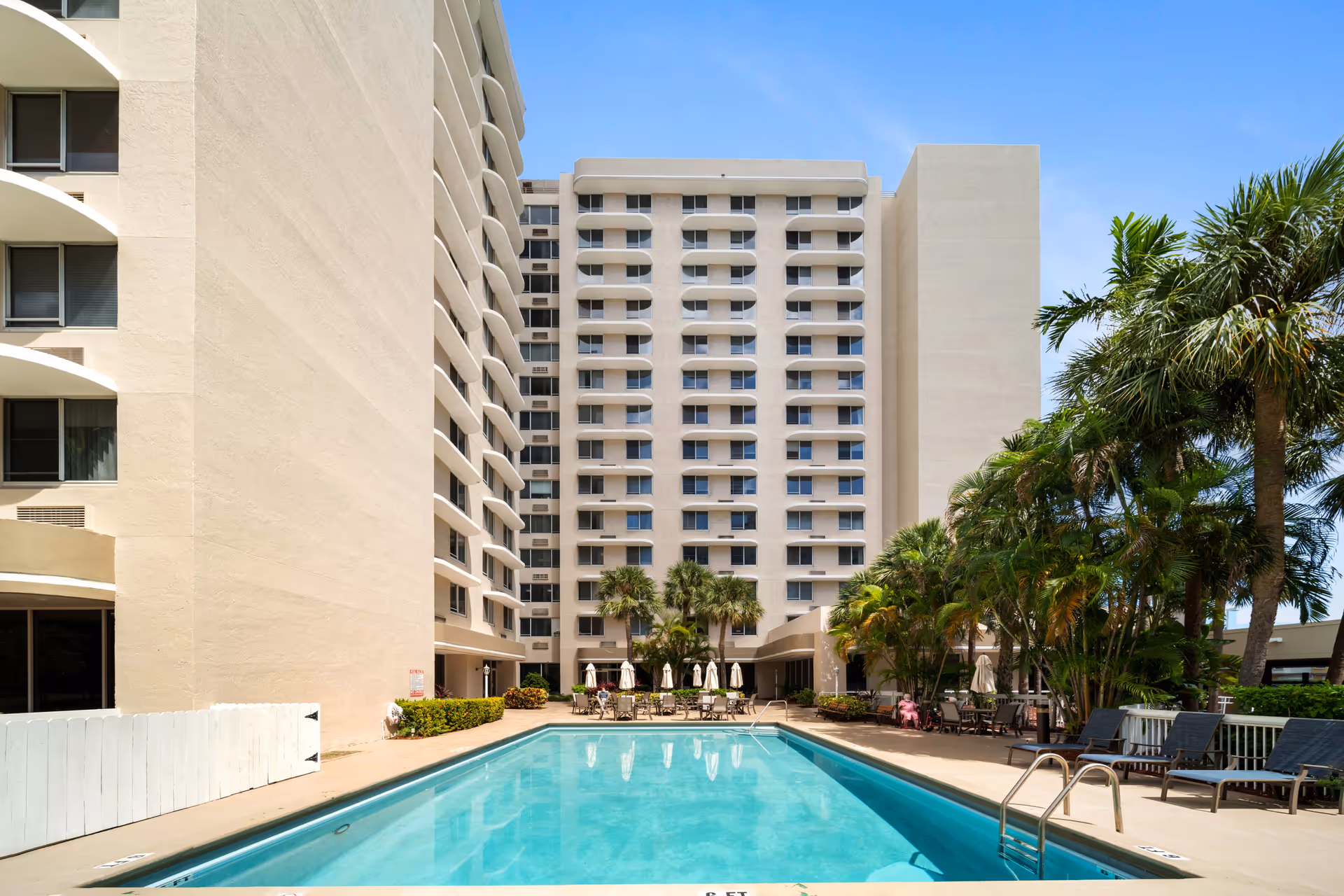 Outdoor swimming pool area at Imperial Club with lounge chairs, tables with umbrellas, palm trees, and a multi-story beige building in the background under a clear blue sky.