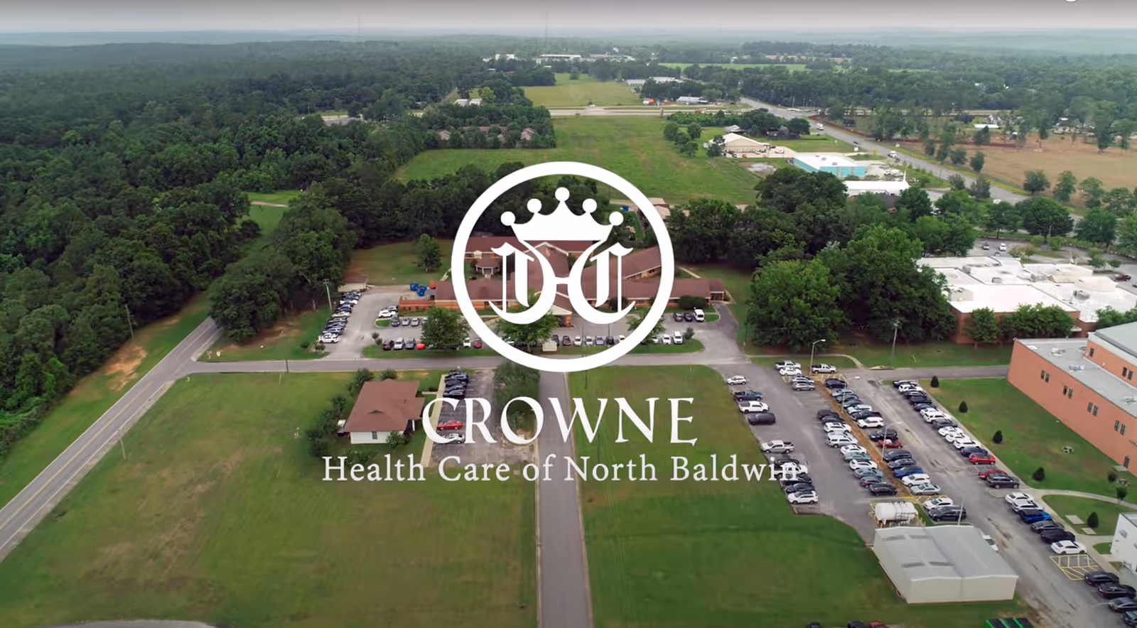 Aerial view of Crowne Health Care of North Baldwin facility showing multiple buildings, parking lots filled with cars, surrounding green fields, and trees.