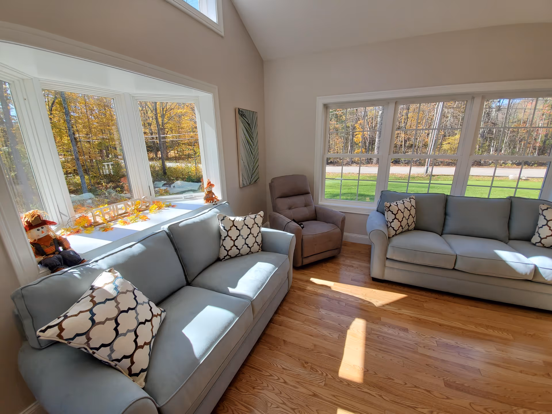 Bright living room with two light-blue sofas, a recliner, large windows showing autumn trees, and hardwood floors.