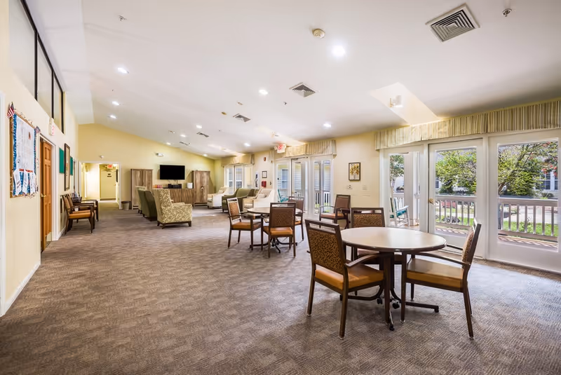A bright memory-care common room with round tables and chairs, lounge seating, and glass doors leading to an outdoor patio.