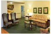 A cozy lounge with a floral sofa, two wingback chairs, a glass-topped coffee table, table lamp, and framed artwork on the wall.
