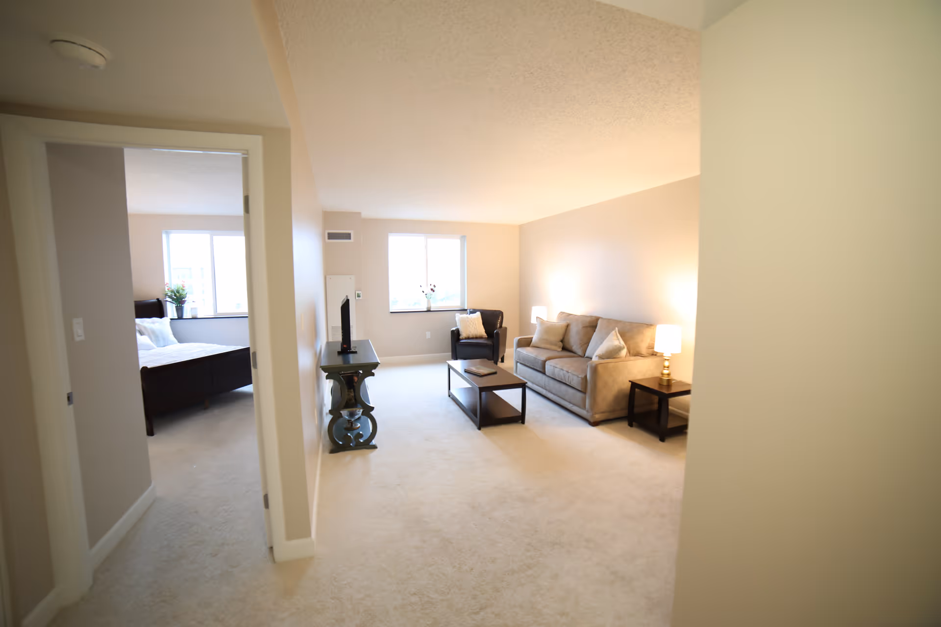 A bright and clean living room with beige walls and carpet. The room features a beige sofa with pillows, a dark armchair, a coffee table, and a side table with a lamp. A TV is placed on a decorative stand against the wall. To the left, an open doorway leads to a bedroom with a bed and a window letting in natural light.