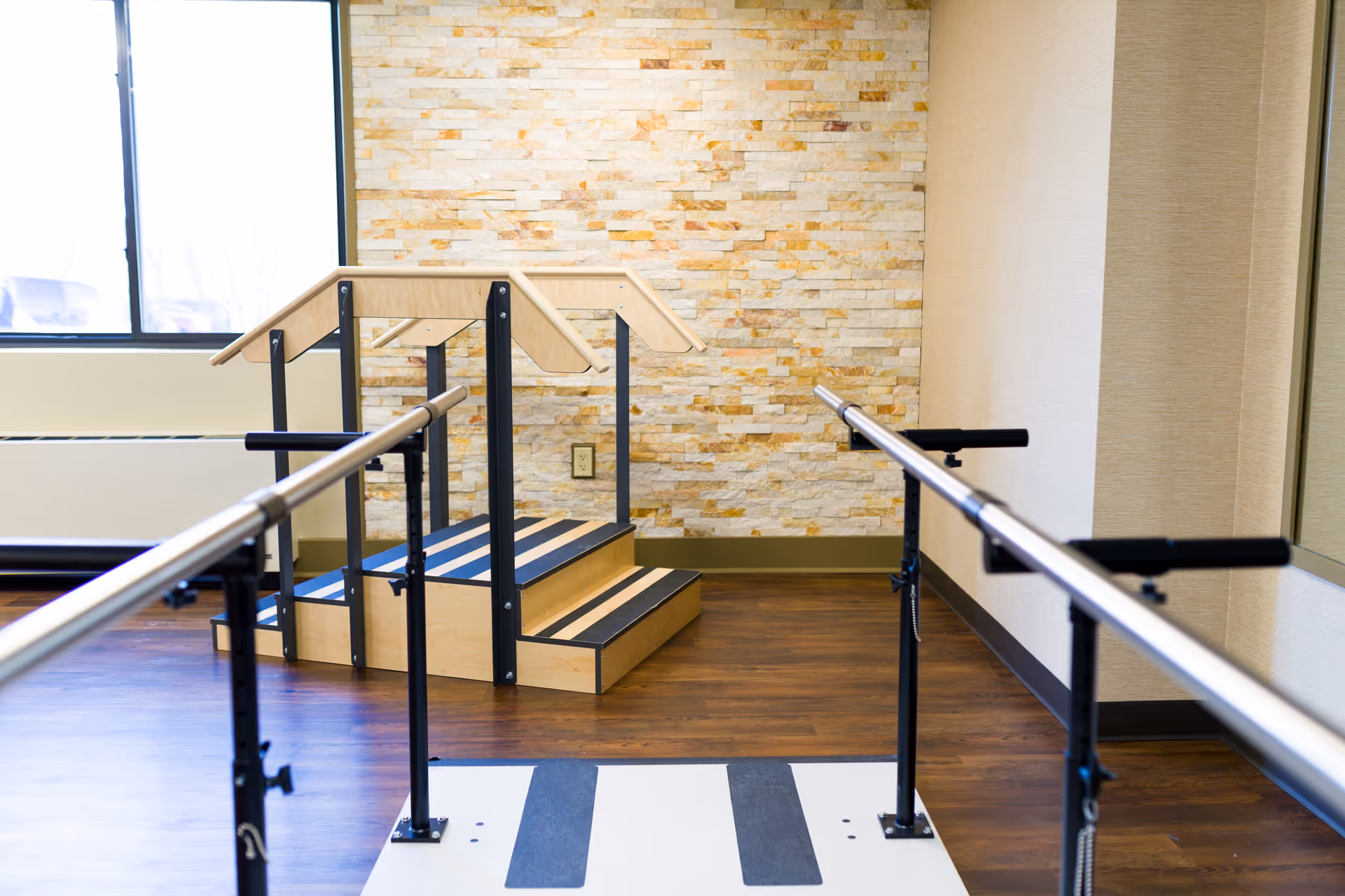 A physical therapy room with parallel bars for walking exercises and a small set of wooden stairs with handrails for rehabilitation training. The room has a stone accent wall, a large window, and wooden flooring.