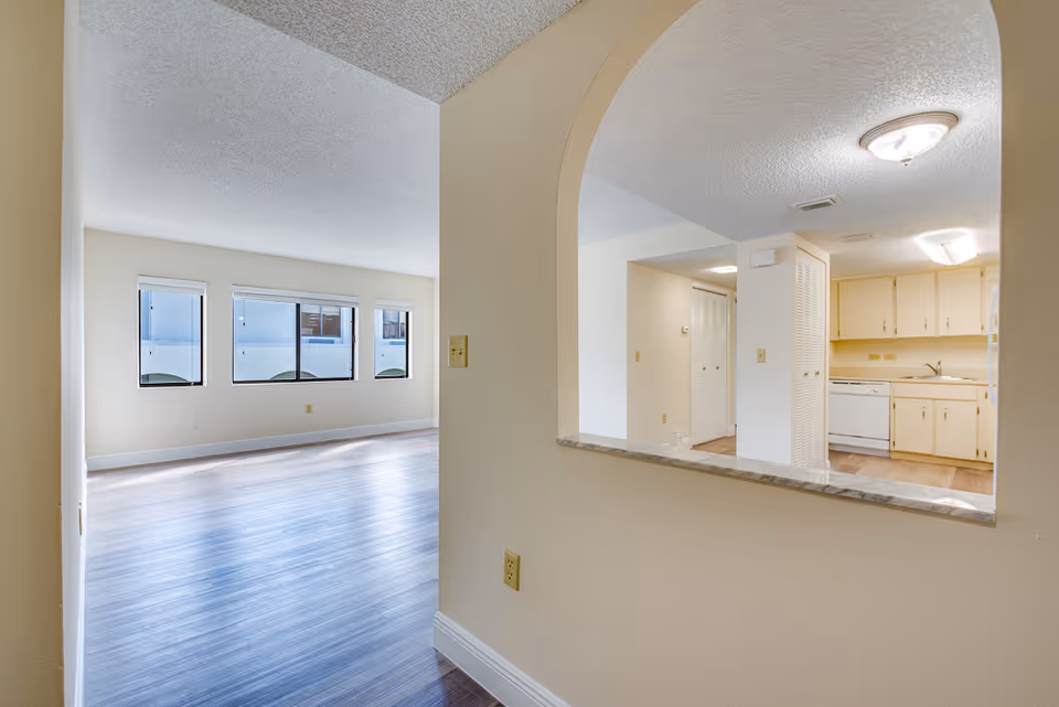 View of an empty living area with three windows letting in natural light and a kitchen visible through an arched pass-through window. The kitchen has light-colored cabinets, a dishwasher, and a sink. The flooring is wood-style laminate throughout.