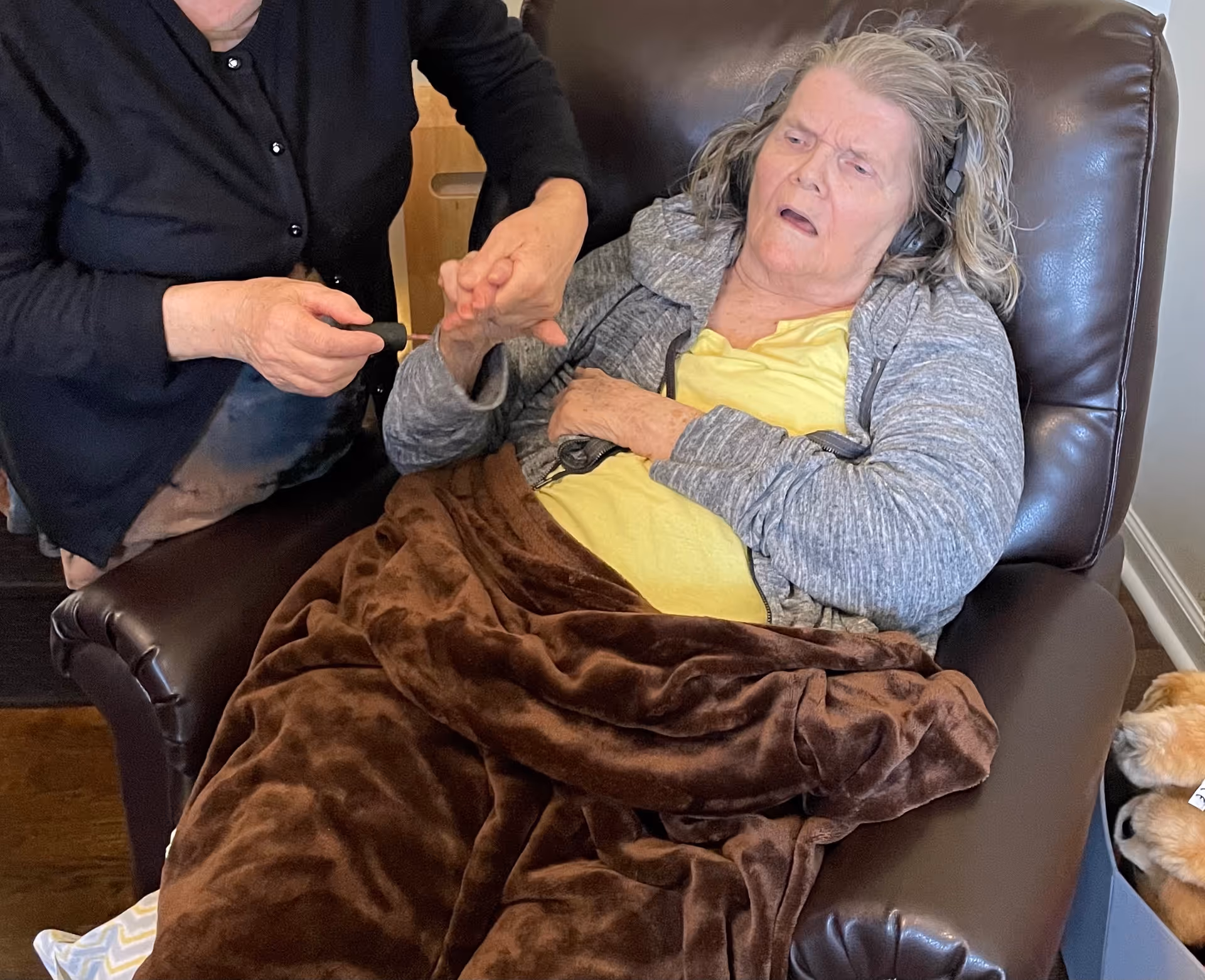 An elderly woman with gray hair wearing headphones and a gray hoodie is sitting in a brown recliner chair covered with a brown blanket. A person in a black cardigan is holding and grooming the elderly woman's fingernails. A small dog is partially visible on the right side of the image.