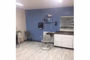 Small interior room with a blue accent wall containing a salon chair, mirror above a white counter, shelving and a wall-mounted dispenser.