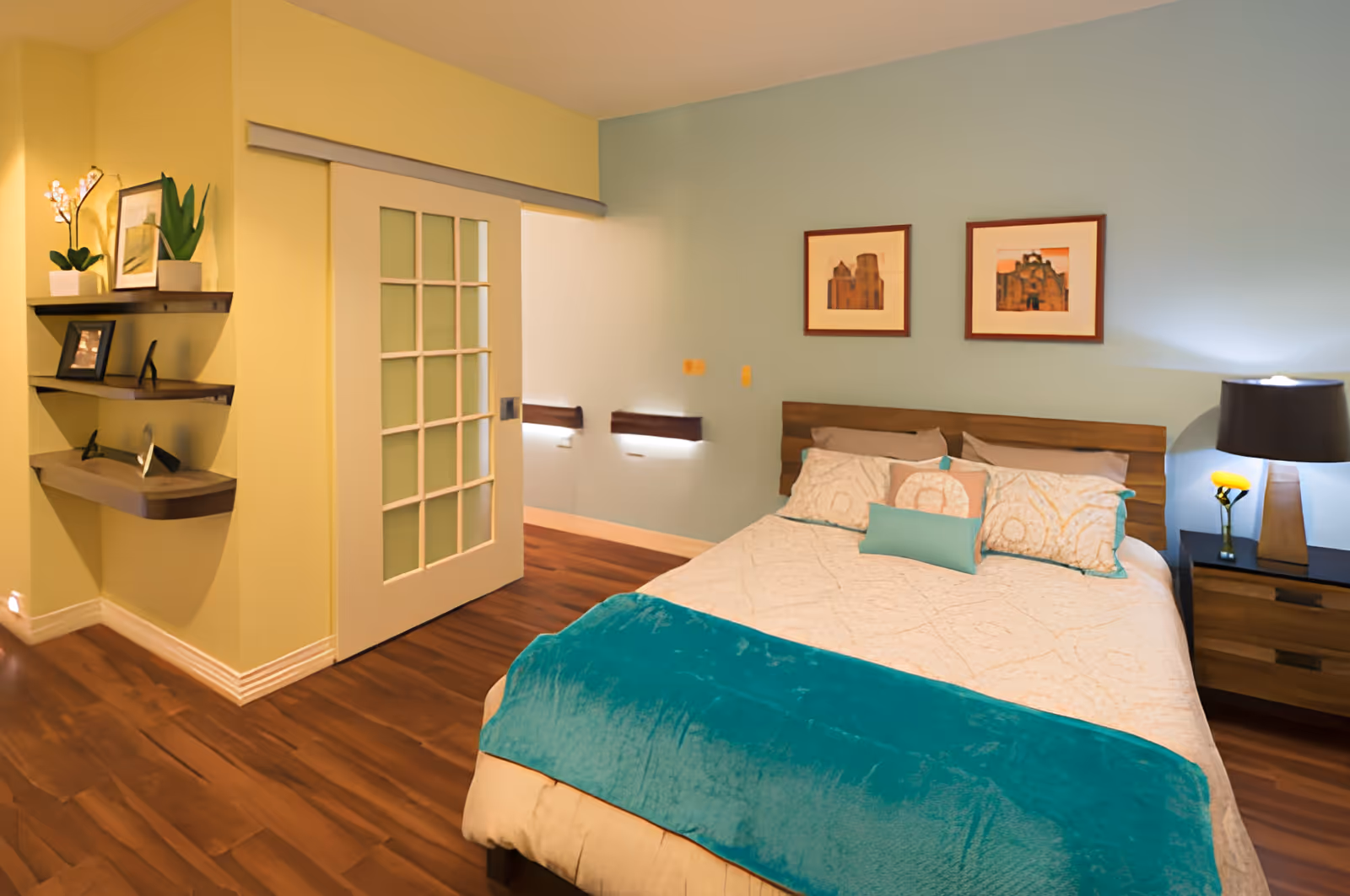 Bright, modern bedroom with a queen bed topped by a teal throw, bedside lamp and nightstand, sliding glass-paneled door, wall shelves and framed artwork.