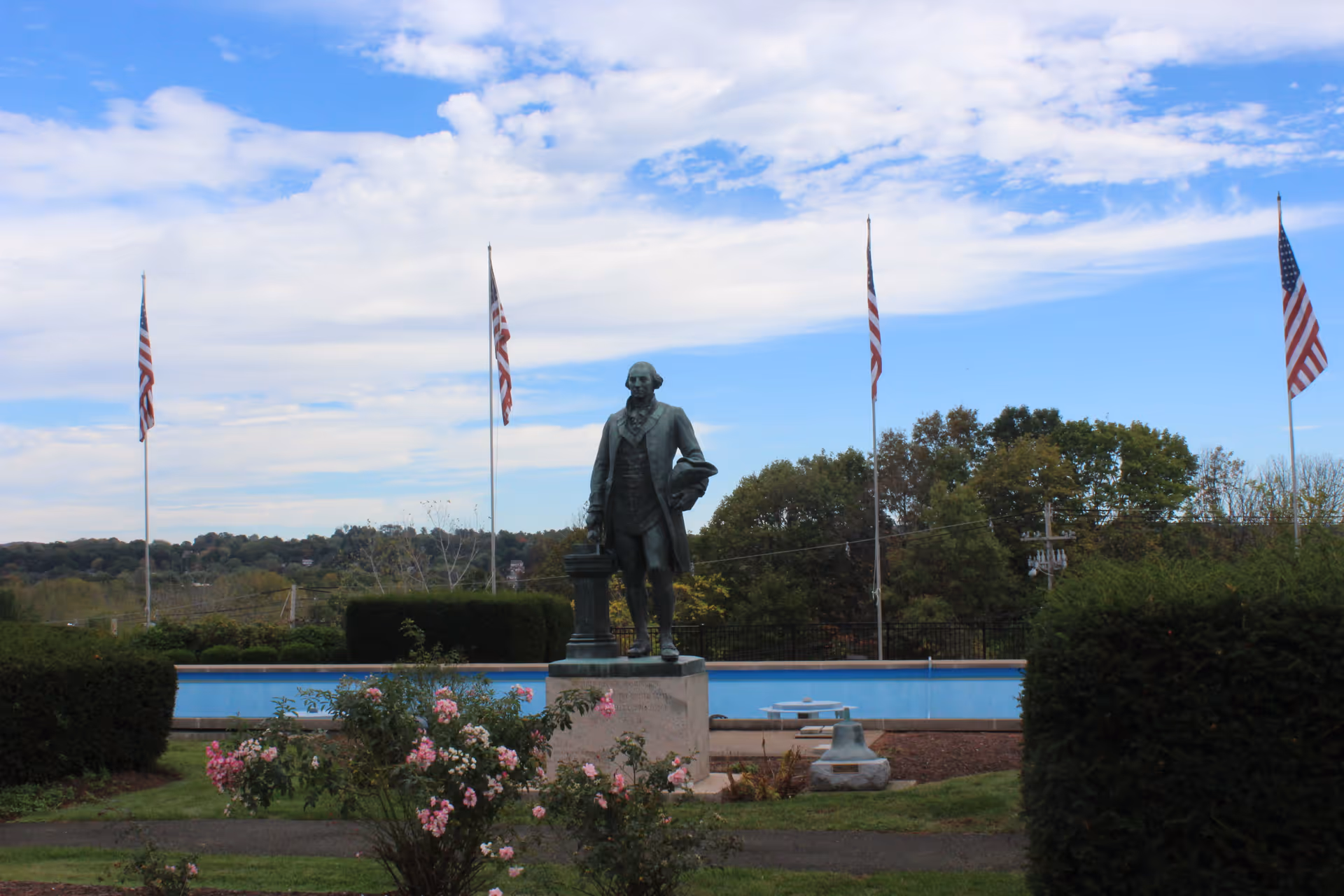 Outdoor garden area with a statue of a man standing on a pedestal surrounded by bushes and pink flowers. Four American flags on tall flagpoles are visible in the background under a partly cloudy sky.