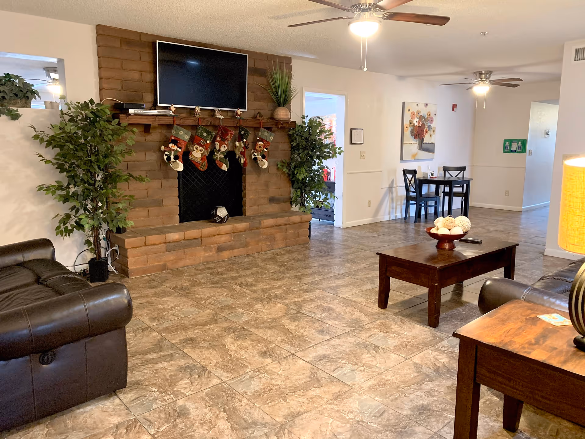 A cozy senior living room with a brick fireplace decorated with Christmas stockings. Above the fireplace is a mounted flat-screen TV. The room has tiled flooring, two leather sofas, a wooden coffee table with decorative balls, and a small dining table with two chairs. There are two ceiling fans with lights, potted plants on either side of the fireplace, and a lamp on a side table.
