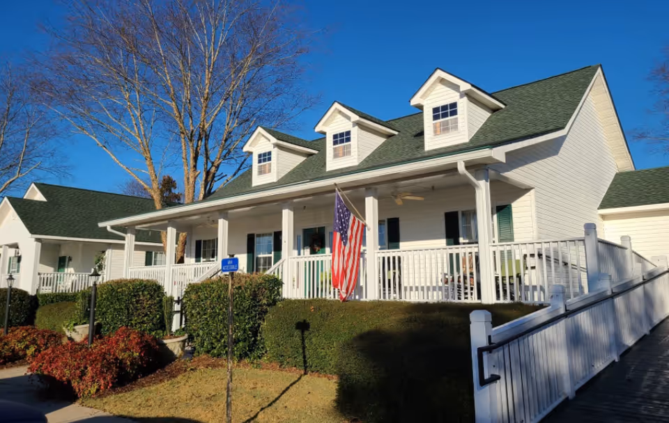 Exterior view of a white building with green roof and three dormer windows, a porch with white railing, an American flag, and surrounding bushes and trees under a clear blue sky.