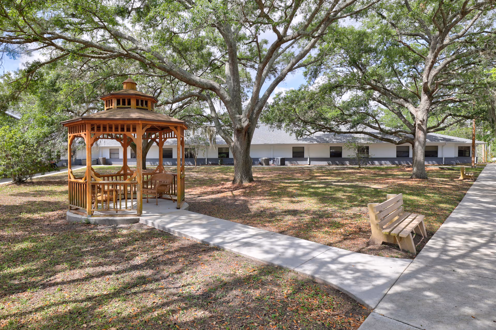 Outdoor garden area at Gardens of Westbrooke featuring a wooden gazebo with seating, large mature trees providing shade, a concrete walkway, and benches along the path. A single-story building is visible in the background.