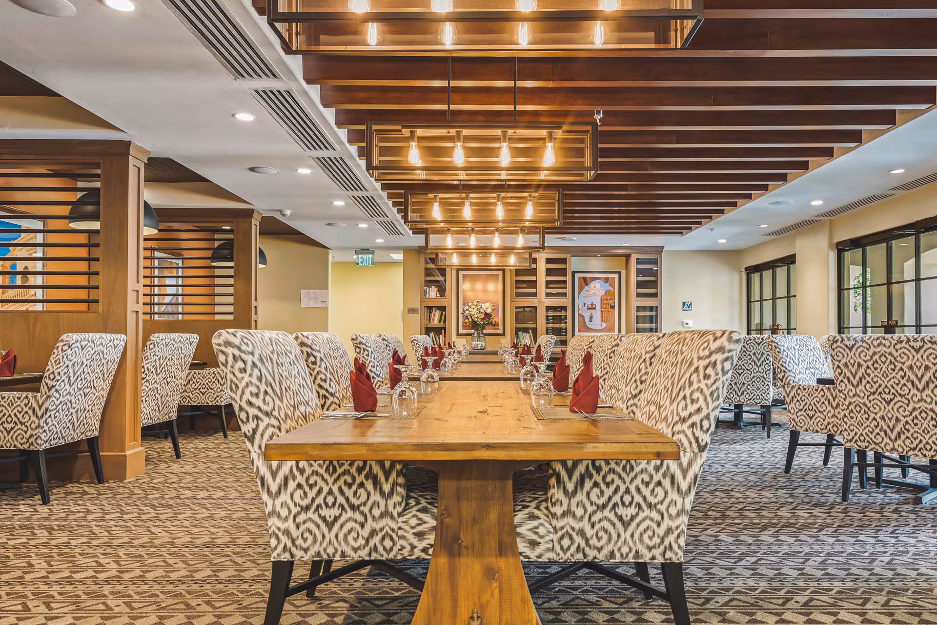 A spacious dining room with a long wooden table set with glasses and red folded napkins. The chairs around the table have a patterned upholstery. The ceiling features wooden beams and modern light fixtures. The room has carpeted floors and large windows on the right side.
