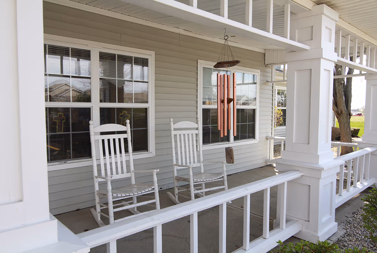 A covered porch area with two white wooden rocking chairs and a hanging copper wind chime. The porch has white railings and columns, and the exterior wall is light gray with two windows reflecting the outside view.