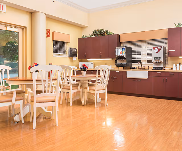 A bright and clean dining area with wooden floors, cream-colored walls, and several white wooden chairs around wooden tables. The back wall features dark brown cabinets, a white farmhouse sink, a coffee machine, and a soda dispenser. There are small potted plants on the tables and above the cabinets, and a glass door leading outside is visible on the left side.