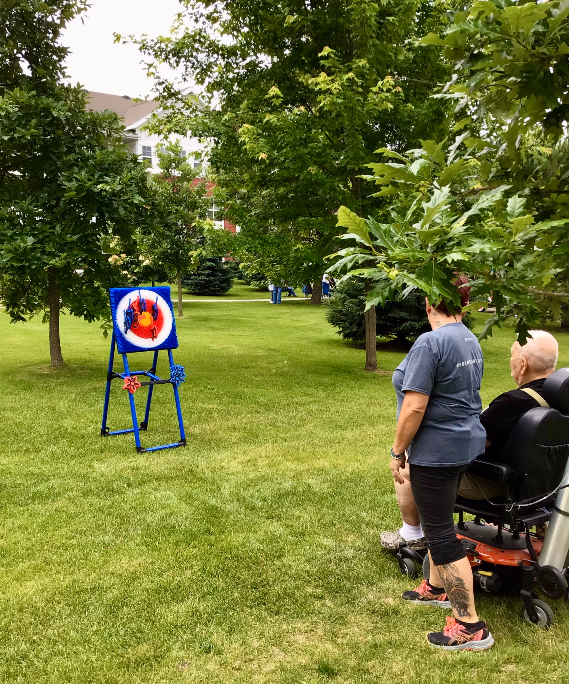 Two people, one standing and one seated in a mobility scooter, play a target toss on a grassy lawn with trees and a building visible in the background.