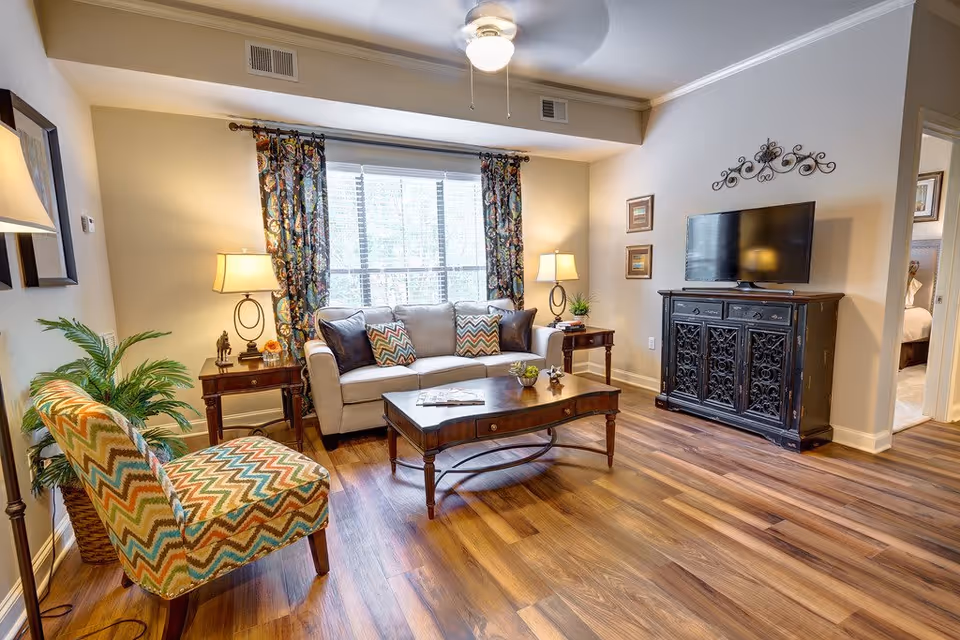 A cozy living room with a beige sofa adorned with patterned and solid pillows, a wooden coffee table, and a colorful zigzag-patterned armchair. There are two side tables with lamps on either side of the sofa, a large window with floral curtains behind the sofa, a ceiling fan with a light, a black ornate TV stand with a flat-screen TV, and wooden flooring throughout. A doorway leads to a bedroom in the background.