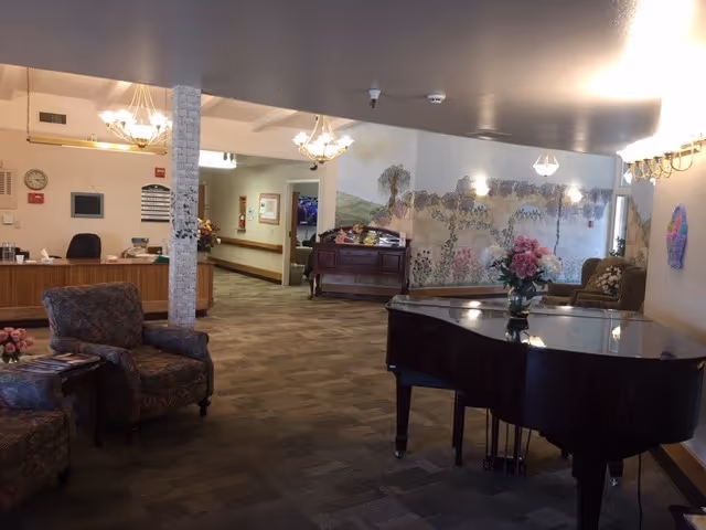 Interior view of a senior living facility lobby area with patterned armchairs, a grand piano with a flower vase on top, a wooden reception desk, chandeliers, and a large mural on the wall depicting a scenic landscape with trees and hills.