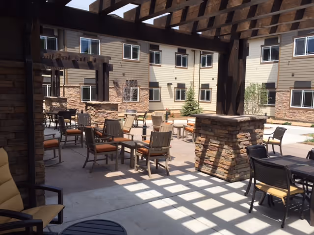 Outdoor patio area with multiple chairs and tables under a wooden pergola, surrounded by a multi-story building with beige siding and stone accents.
