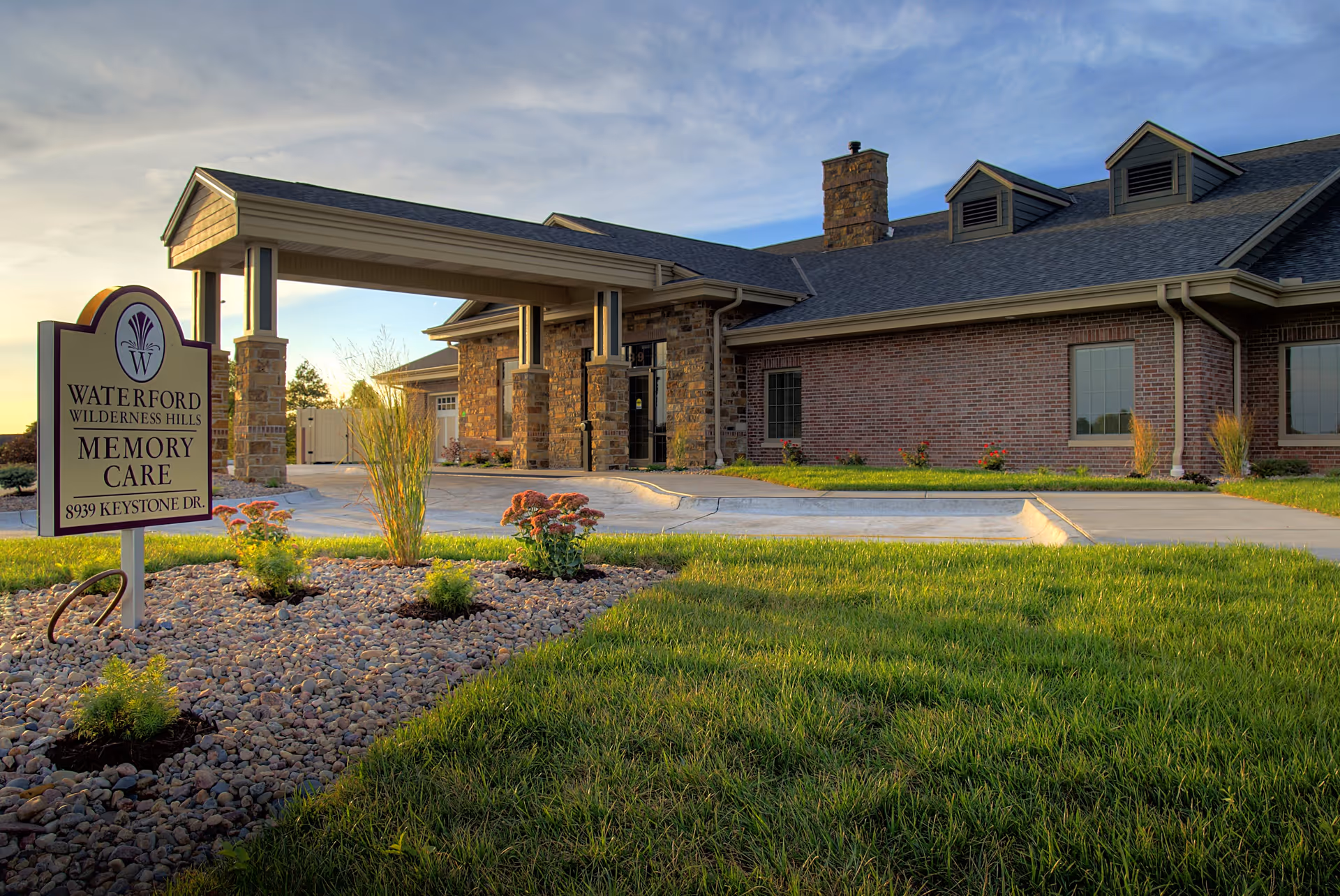 Exterior view of Waterford at Wilderness Hills Memory Care facility showing a brick and stone building with a covered entrance, landscaped garden with grass, flowers, and rocks, and a sign displaying the facility name and address.