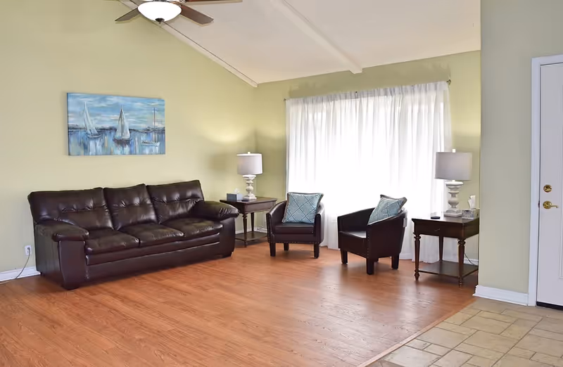 Bright living room with a dark leather sofa, two armchairs, side tables and lamps in front of a curtained window.