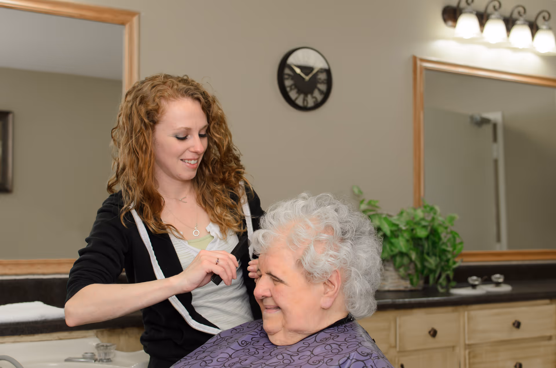A young woman with curly hair is smiling and cutting the hair of an elderly woman with white curly hair in a salon-like setting with mirrors, a clock on the wall, and a plant on the counter.