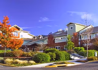 Front exterior of a senior living building with landscaped shrubs, autumn trees, a walkway and entrances under a blue sky.