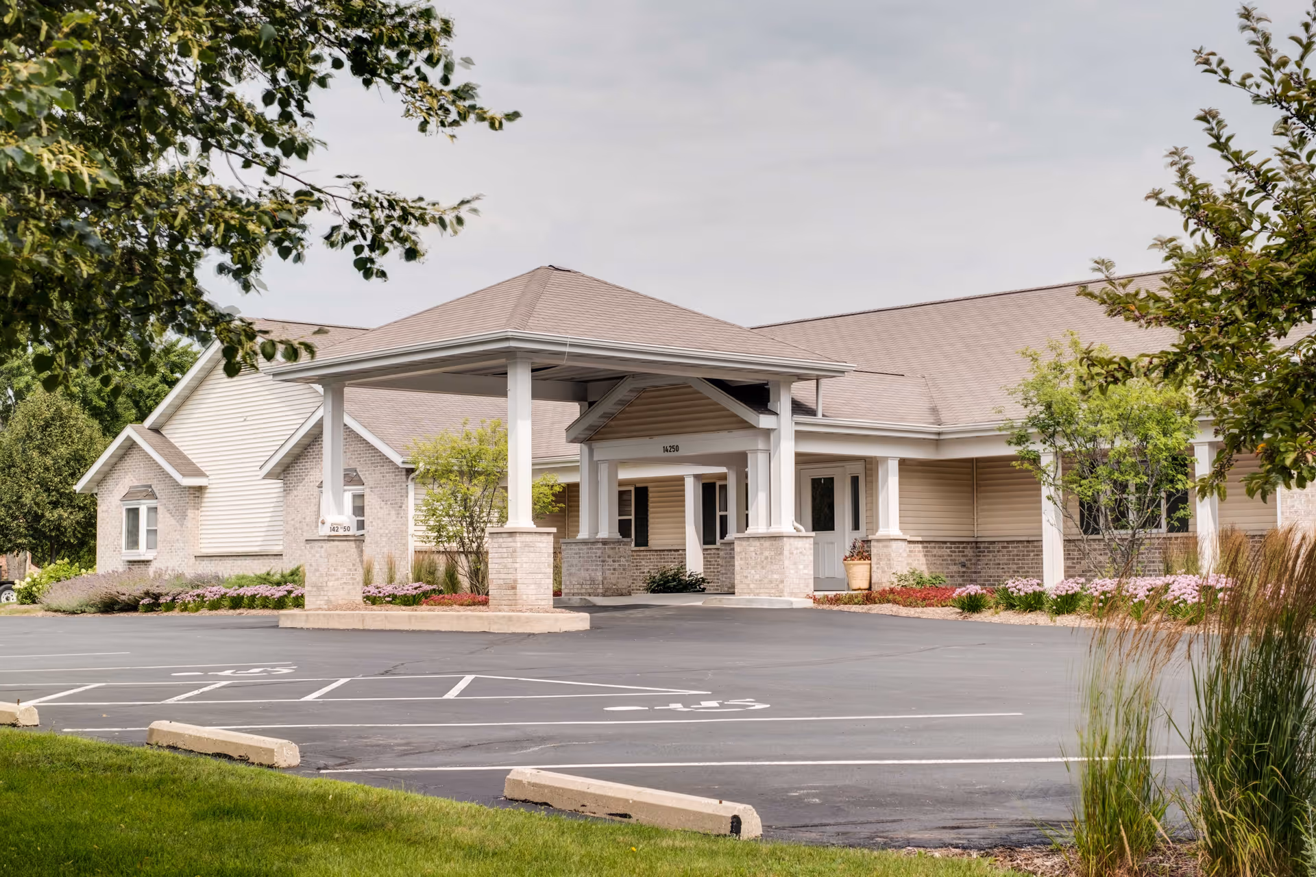 Exterior view of a single-story senior living facility building with a covered entrance, surrounded by landscaped greenery and a parking lot with marked spaces.