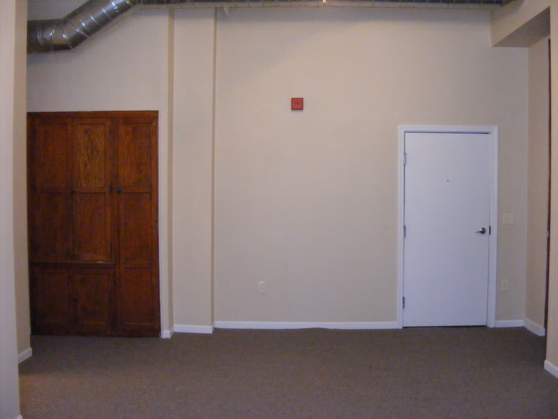 An empty interior room with beige walls and carpeted floor. There is a white door on the right side and a wooden cabinet with multiple doors on the left side. A red fire alarm is mounted on the wall above an electrical outlet.