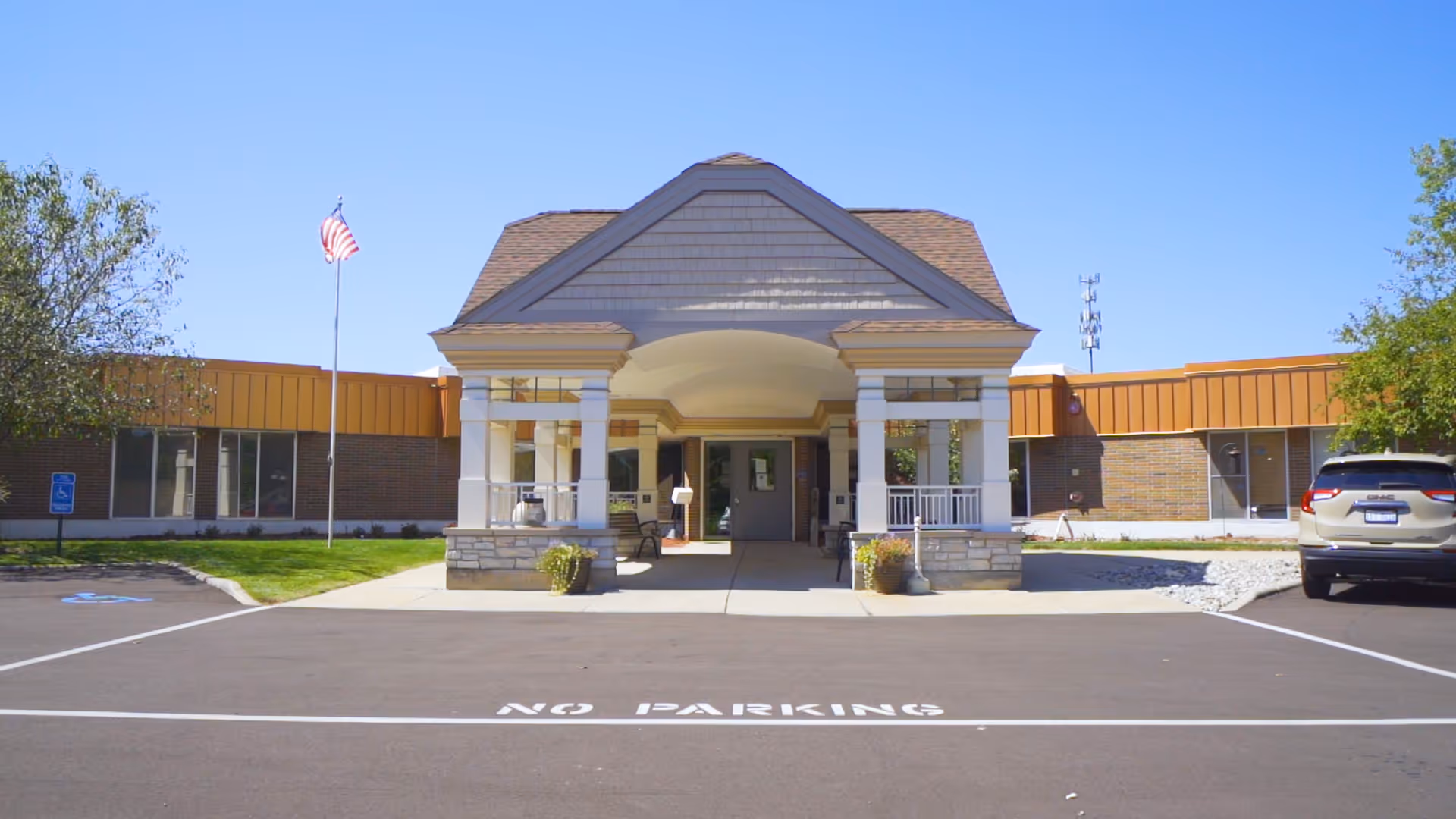 Front exterior view of Medilodge of Lansing building with a covered entrance supported by white columns, an American flag on a flagpole to the left, a parked car to the right, and clear blue sky above.
