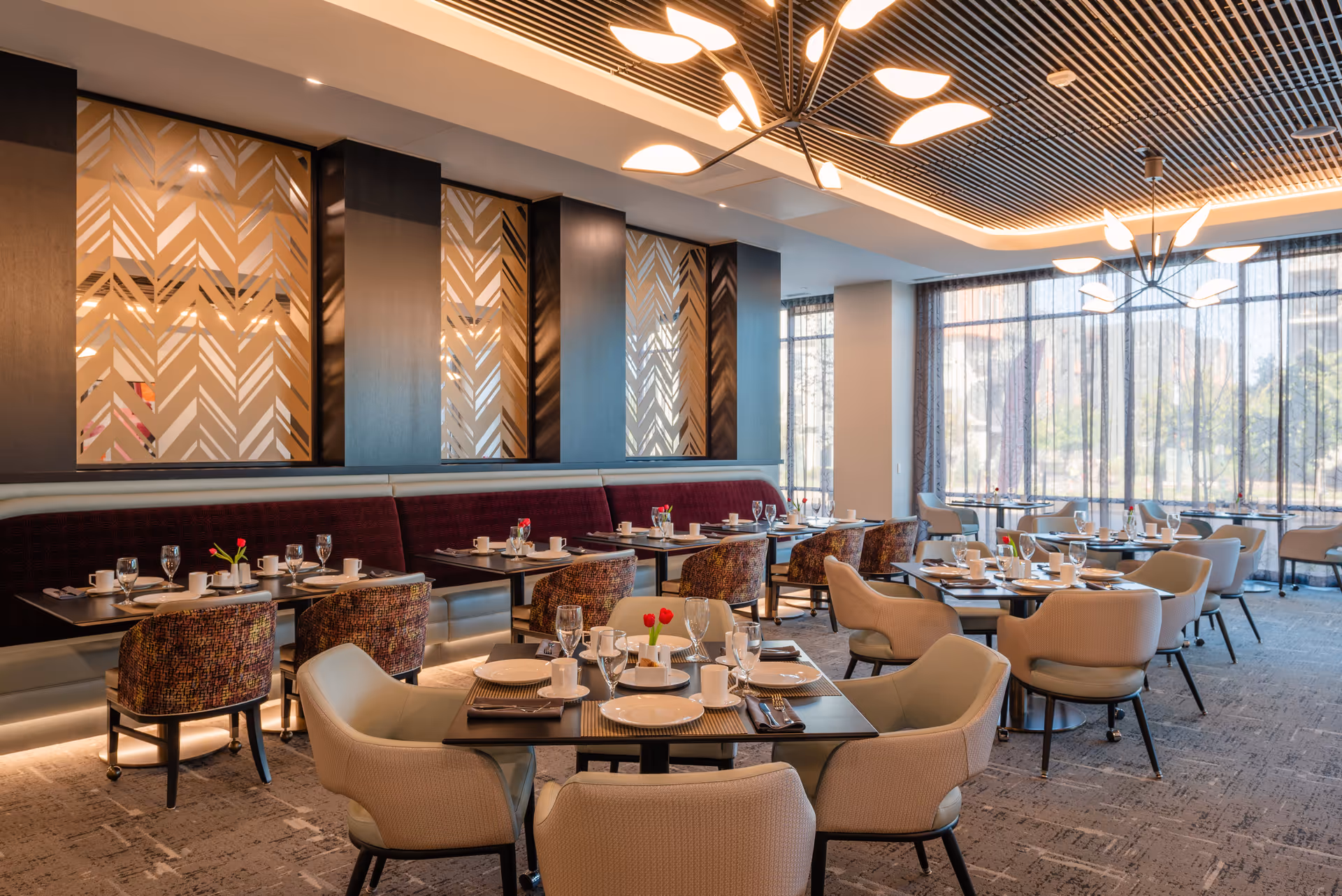 A modern dining room with several tables set with white plates, cups, glasses, and silverware. The room features upholstered chairs in beige and patterned fabric, a long maroon banquette along the wall, decorative wall panels with geometric patterns, large windows with sheer curtains, and contemporary ceiling light fixtures.
