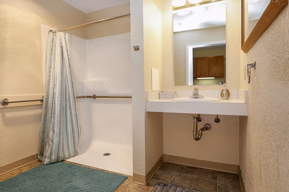 A bathroom with a walk-in shower featuring a striped shower curtain and grab bars. There is a sink with a large mirror above it, a soap dispenser, and a small stack of folded towels. The floor is tiled and there is a blue bath mat in front of the shower.