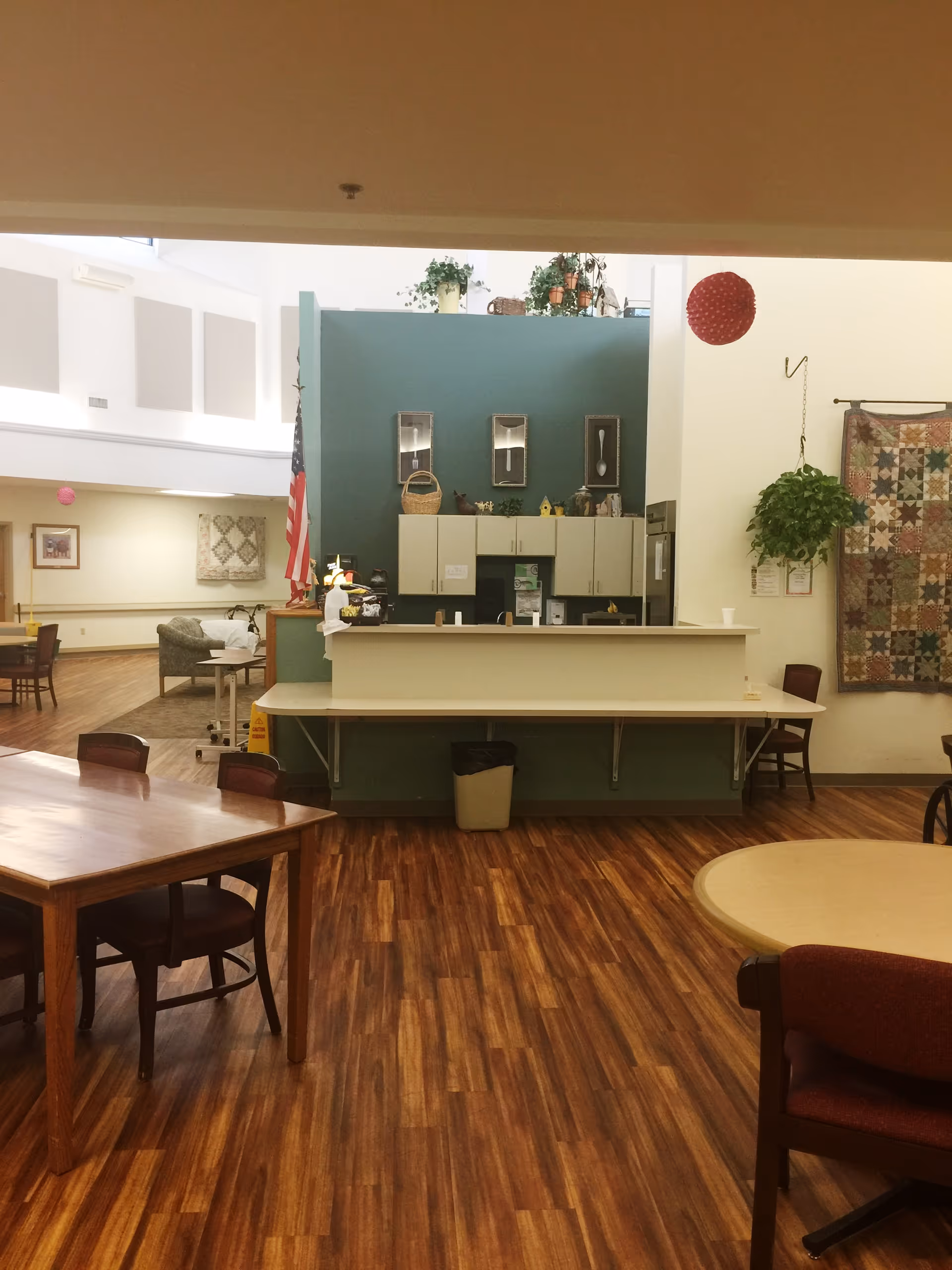 Open common area with wood floors, tables and chairs and a central service counter/kitchen decorated with plants and a quilt on the wall.