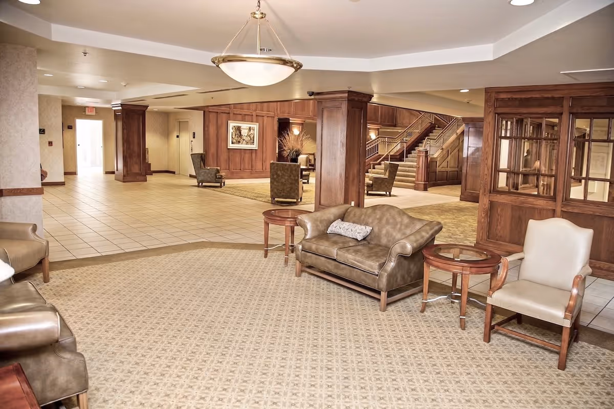 A spacious senior living facility lobby with beige patterned carpet and tiled floor. The area features brown leather sofas, beige armchairs, wooden side tables, and wood-paneled walls. A large light fixture hangs from the ceiling, and a staircase with wooden railings is visible in the background.