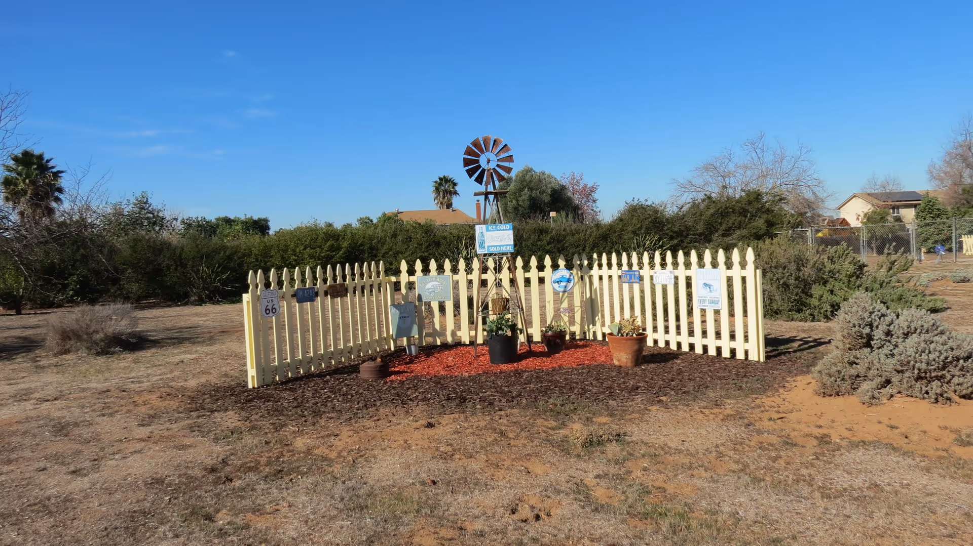 A small fenced garden area with a yellow picket fence surrounding a decorative windmill and several potted plants. Various vintage-style signs are attached to the fence, including one that says 'Ice Cold Coca-Cola Sold Here'. The garden is set in a dry, grassy outdoor area with bushes and trees in the background under a clear blue sky.