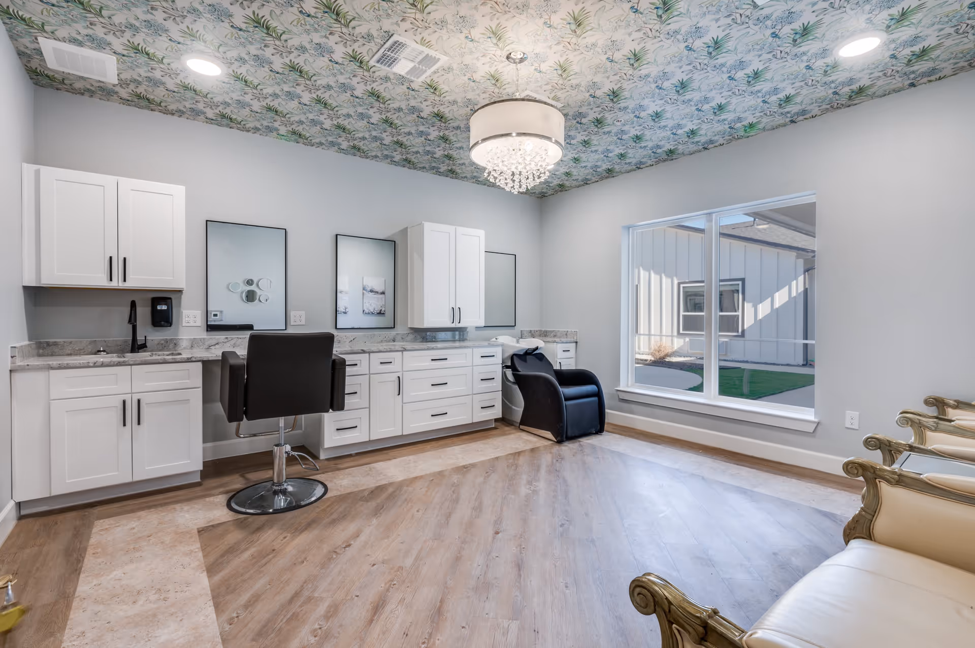 A bright and modern salon room with white cabinetry, a black salon chair in front of two mirrors, a black shampoo chair, and a large window letting in natural light. The ceiling has a decorative floral pattern and a chandelier light fixture. There are also ornate cream-colored chairs along one wall.