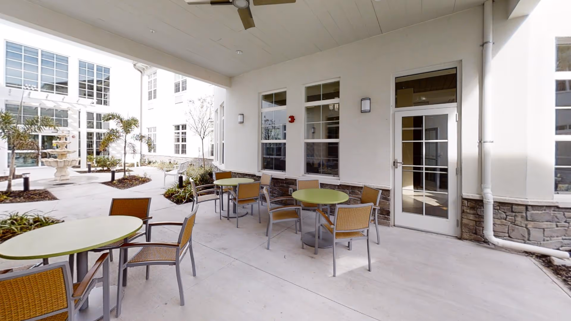 Covered outdoor patio area with several round tables and chairs. The patio is adjacent to a building with large windows and a glass door. In the background, there is a courtyard with a fountain, small palm trees, and landscaped garden beds.