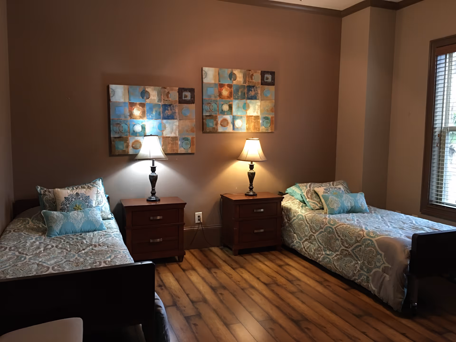 A cozy bedroom with two single beds, each with patterned bedding and multiple pillows. Between the beds are two wooden nightstands, each with a table lamp. Above the nightstands are two abstract paintings with blue, brown, and beige tones. The room has wooden flooring and a window with blinds on the right side.