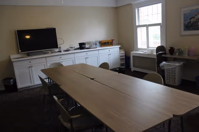 A meeting or activity room with a long rectangular table made of two tables pushed together, surrounded by chairs. Against the wall is a white cabinet with a flat-screen TV on top, along with some glassware and other items. A window on the right side lets in natural light, and there is a laundry basket and some additional items near the window.