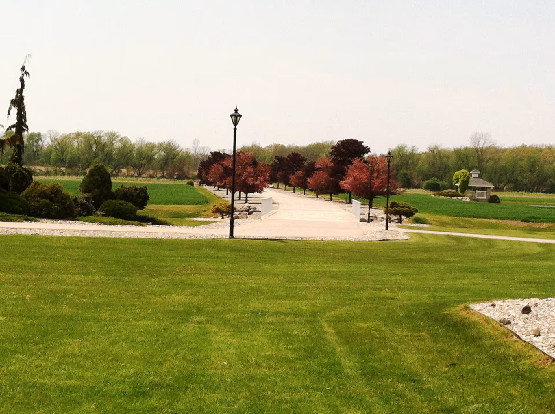 A wide green lawn leading to a paved, tree-lined walkway with lampposts and a small gazebo in the distance.