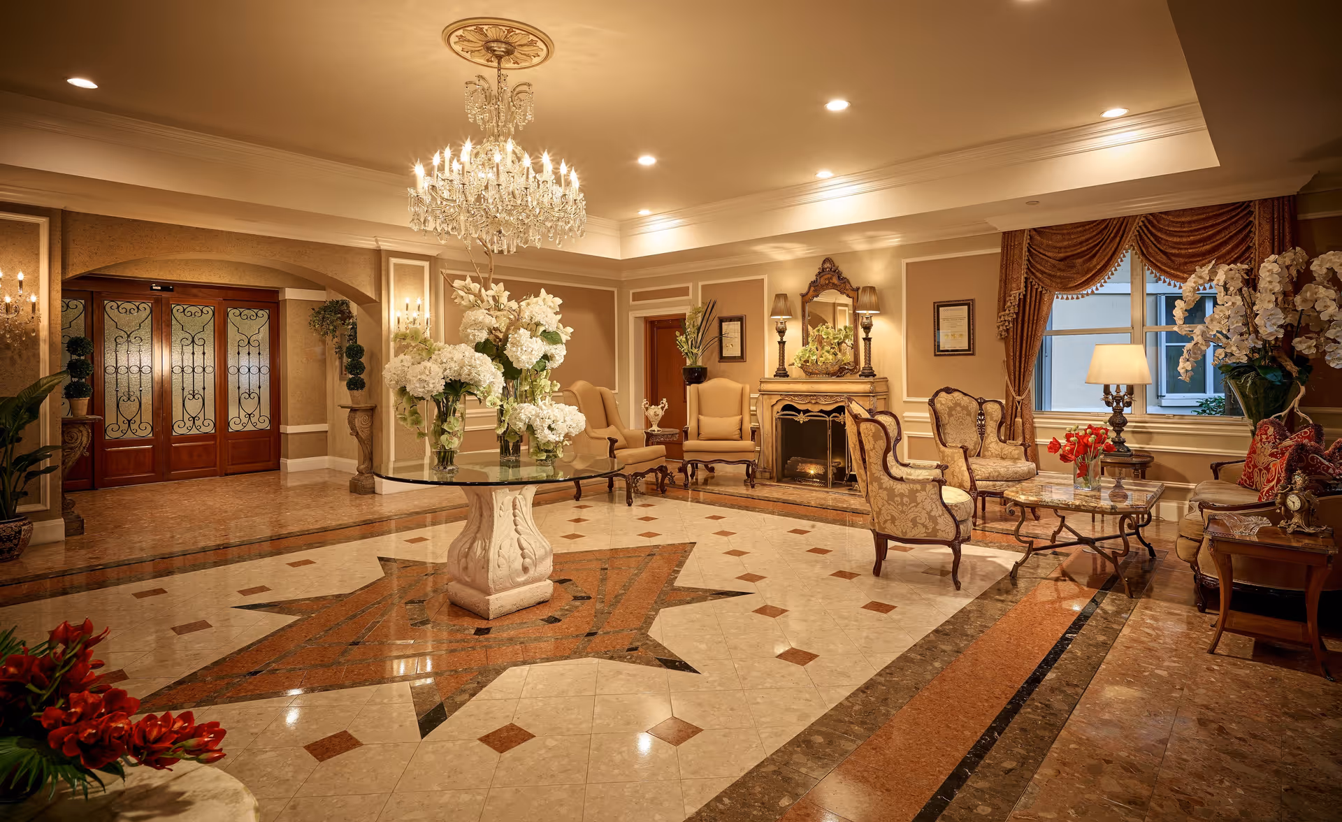 Luxurious interior lobby with a central glass table topped with flowers, chandelier, marble floors, and upholstered seating around a fireplace.