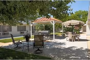 Outdoor patio area with a covered gazebo, metal chairs, a grill, and a table with an umbrella surrounded by a building and green lawn under a tree providing shade.