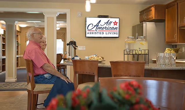 Two elderly residents sitting at a kitchen counter in a communal dining area with an 'All American Assisted Living' sign on the wall.
