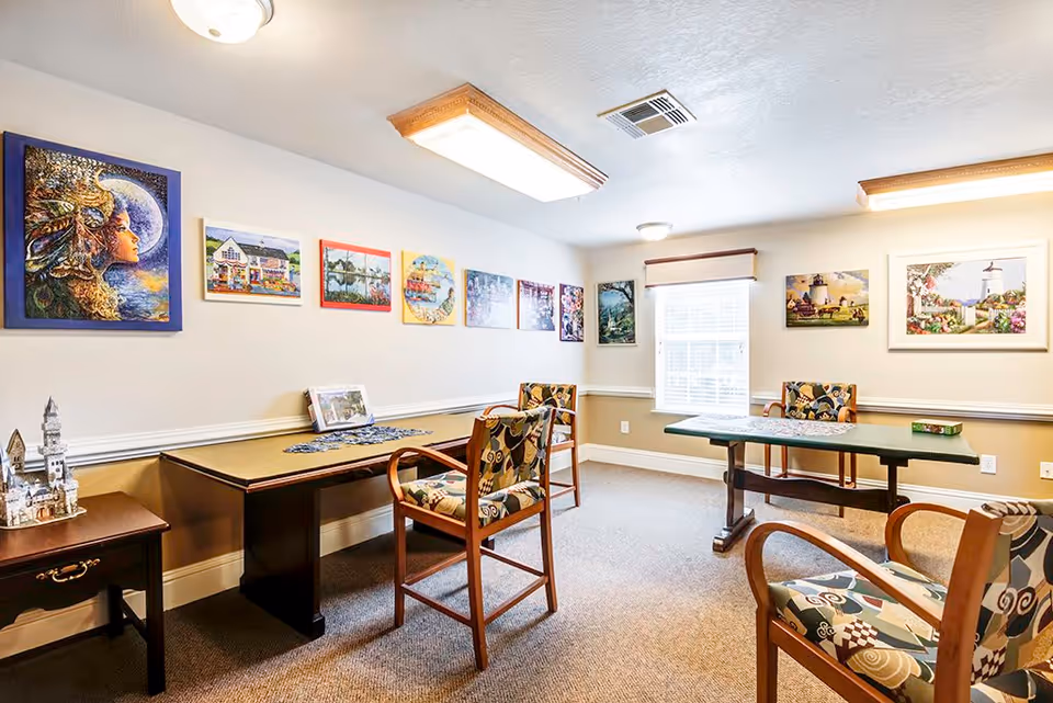 A bright room with two tables and several patterned chairs arranged around them. The walls are decorated with colorful paintings and artwork. A window with blinds allows natural light to enter the room. The carpeted floor and ceiling lights create a cozy atmosphere.