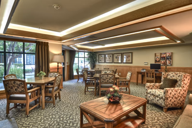 A well-lit common area in a senior living facility featuring multiple wooden tables and chairs with floral upholstery, a patterned carpet, large windows with curtains allowing natural light, framed artwork on the walls, a computer desk with a chair, and a wooden coffee table with a floral centerpiece.