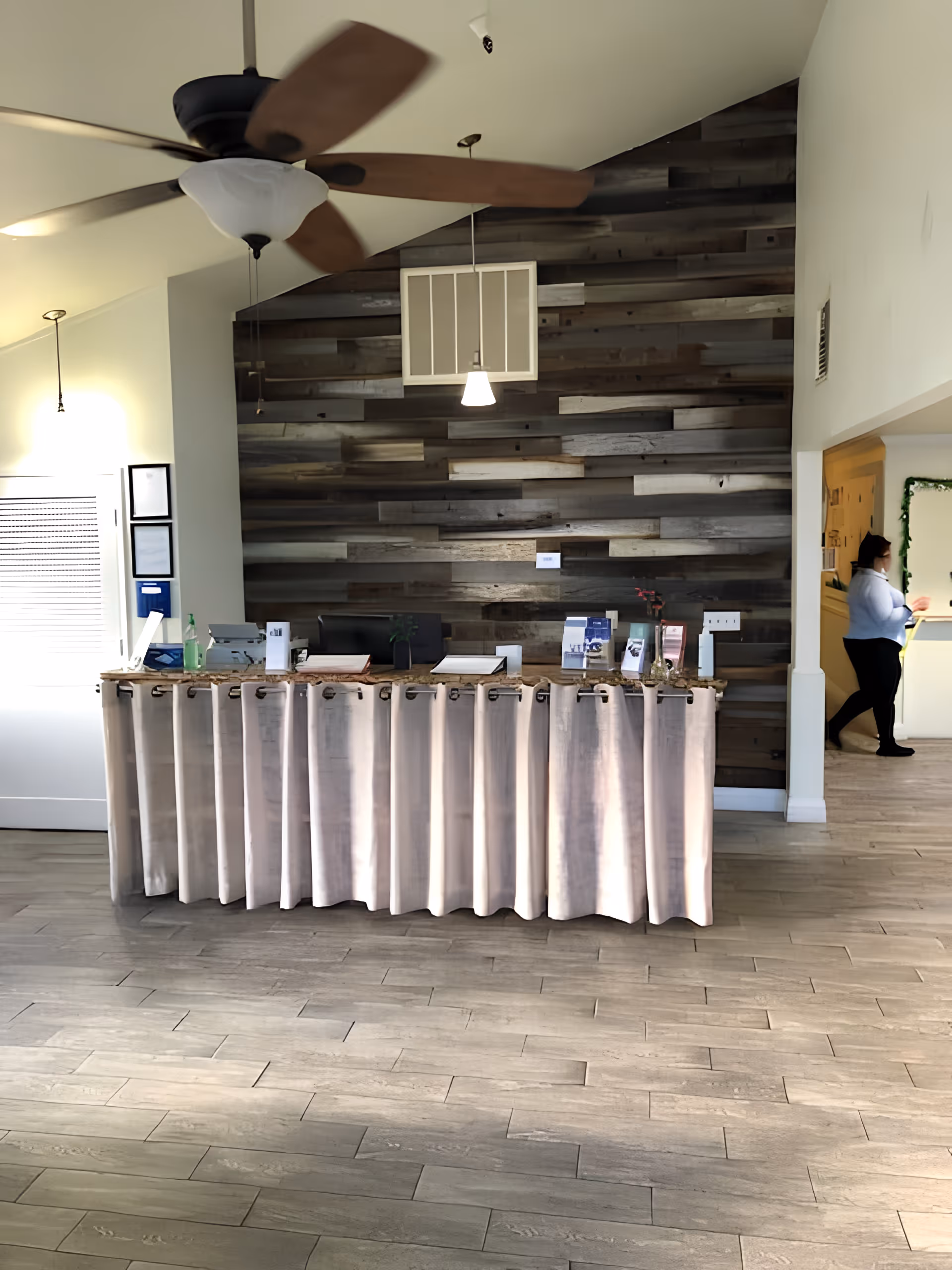 Reception area of Solano Life House Assisted Living & Memory Care with a wooden paneled wall behind a counter draped with a light-colored curtain. A ceiling fan with wooden blades and a light fixture hangs from the ceiling. A person is walking in the hallway to the right.