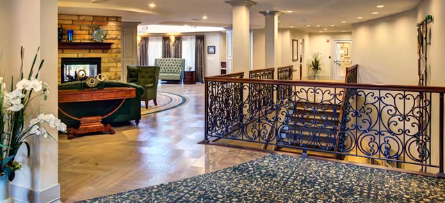 Interior view of a senior living facility featuring a carpeted area with floral patterns in the foreground, a decorative wrought iron railing along a staircase, a wooden table with a green upholstered chair, a stone fireplace, and additional seating with armchairs and a sofa in the background. The space is well-lit with recessed ceiling lights and decorated with plants and framed artwork.
